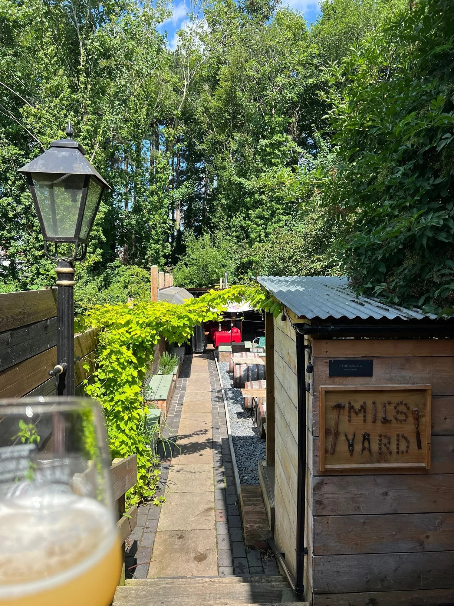 Photograph of an outdoor beer garden named "Mils Yard" featuring wooden structures, a pathway lined with greenery, and tables with red chairs in the background. A glass of beer is partially visible in the foreground, with tall trees and a clear blue sky overhead.