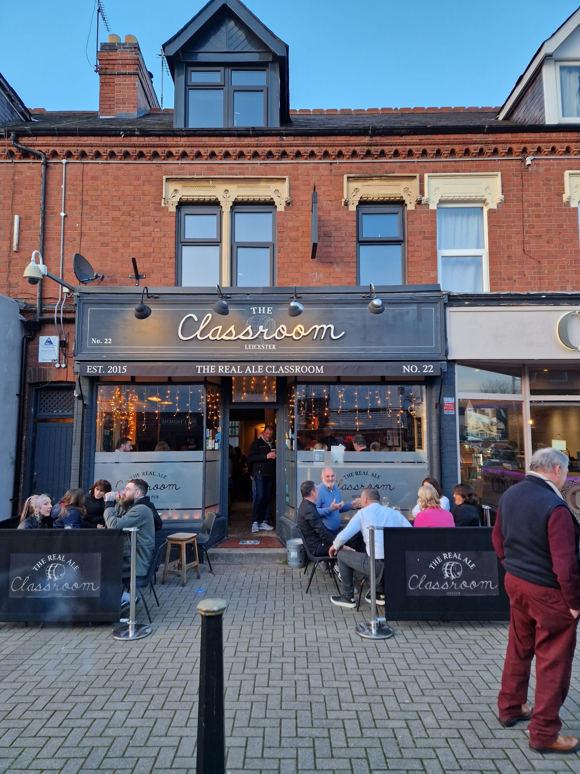Photograph of a street view featuring a brick building with a black storefront sign reading "The Classroom" and outdoor seating area with people dining. The scene includes string lights inside the establishment, black sandwich boards with white text, and a clear blue sky overhead.