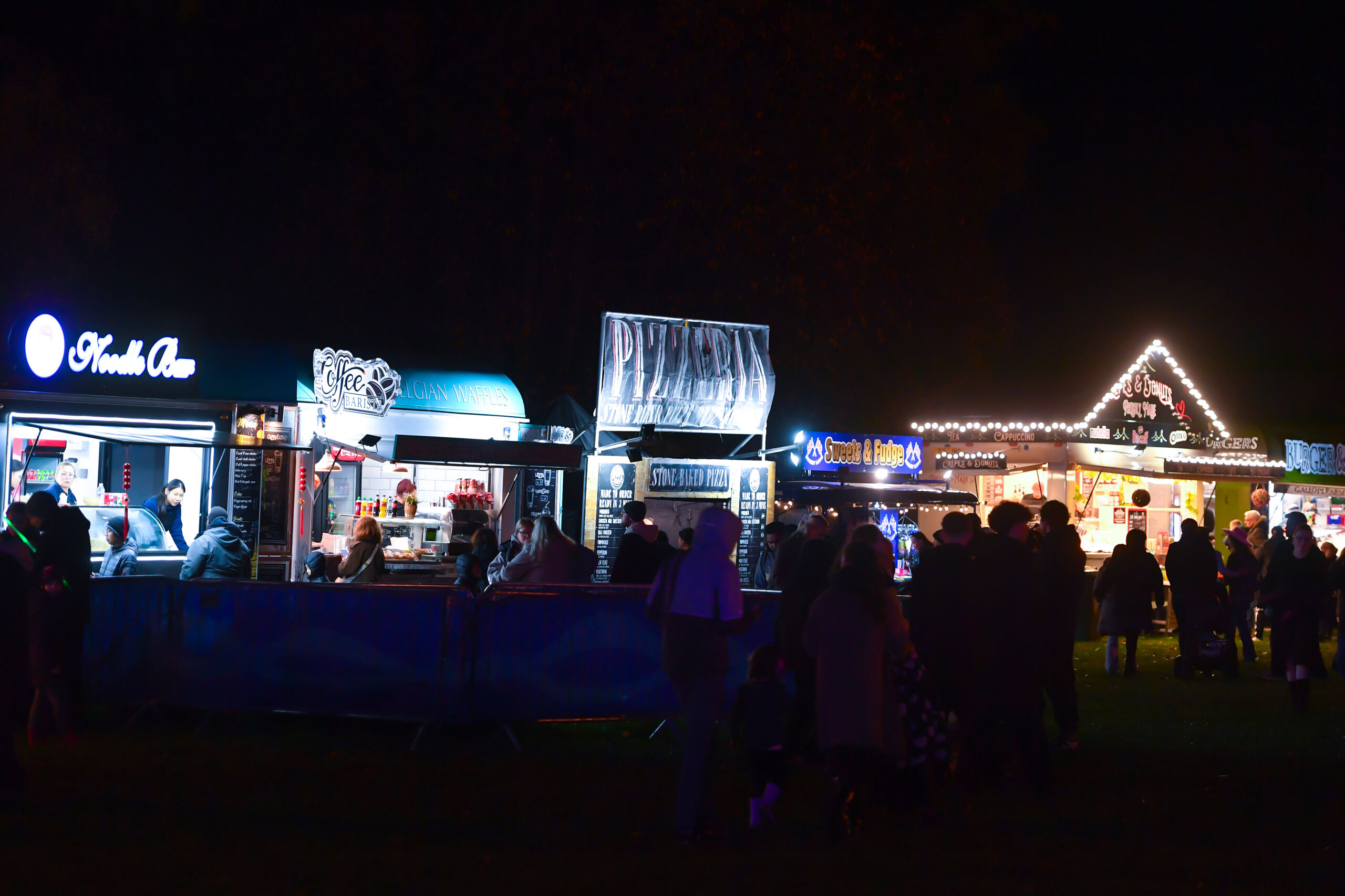 Photograph of a nighttime outdoor food market featuring several illuminated vendor stalls with signs, including "French Oven" and "Pizzas." Crowds of people are gathered in front of stalls, with one building decorated with string lights and a triangular roof adorned with holiday lights.