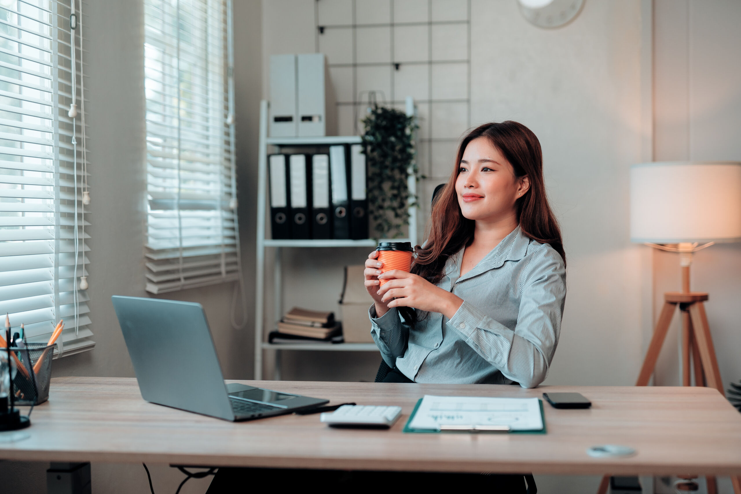 Photograph of a woman sitting at a desk in a modern office holding a cup, with a laptop, clipboard, and smartphone on the desk. Background includes shelves with binders, a plant, a wall clock, and a floor lamp, suggesting a professional work environment.