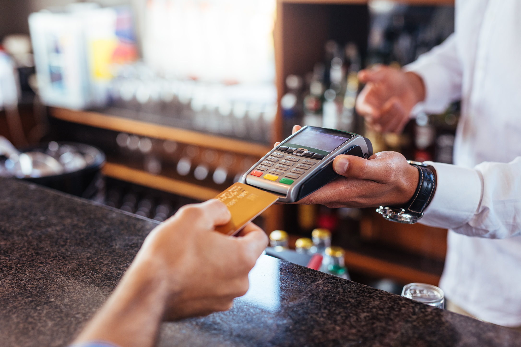 Photograph showing contactless card payment at a bar counter, with customer presenting a bank card to handheld payment terminal. Bartender holds terminal beside drinks shelves and bottles, highlighting quick electronic transaction in hospitality setting.