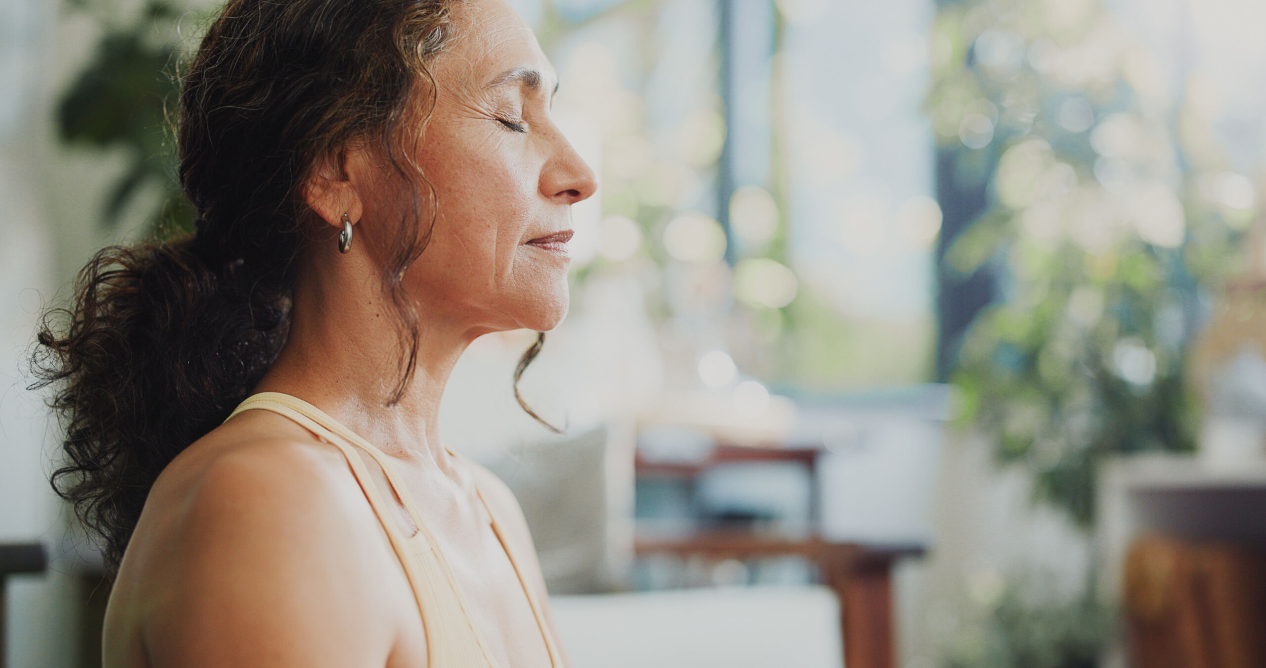 Photograph of a woman with closed eyes, appearing calm and meditative in a softly lit indoor setting. Background includes blurred greenery and furniture, suggesting a peaceful, home environment focused on relaxation or mindfulness.