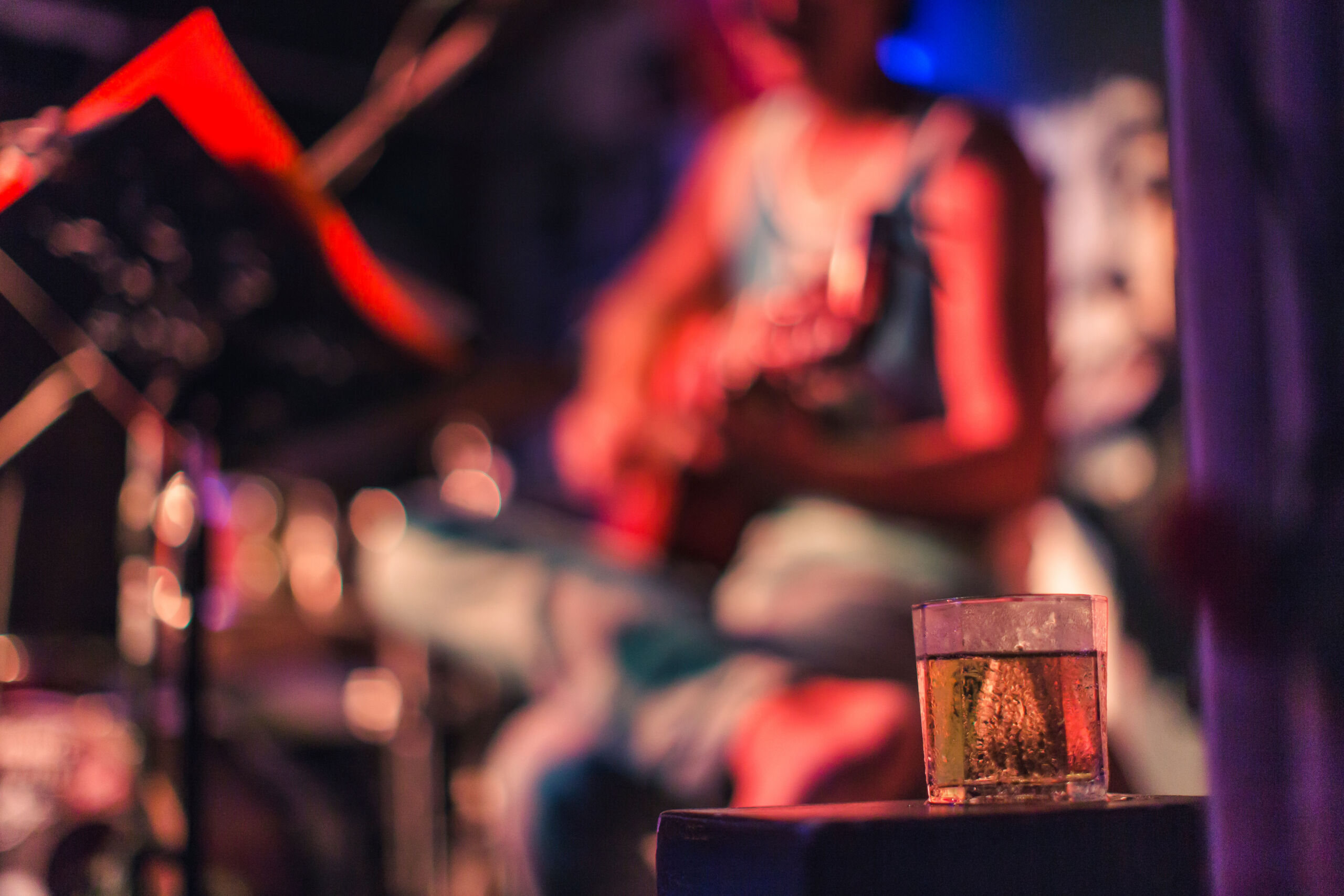 Photograph captures a blurred live music performance, with guitarist and drum kit under red and blue stage lighting. A half-filled glass sits sharply in focus on a foreground speaker, contrasting with indistinct performers and equipment behind.