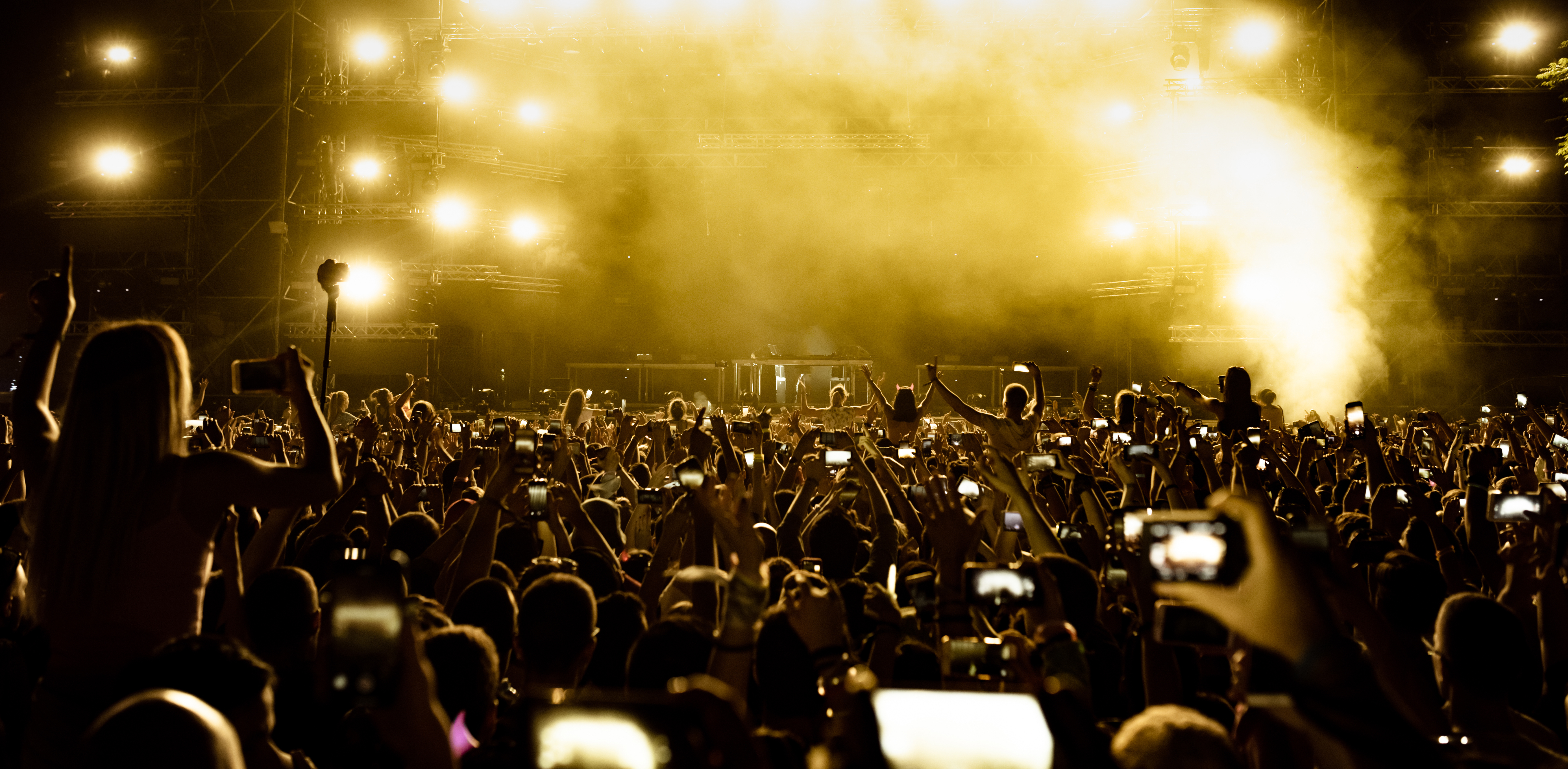 Photograph of a large crowd at a concert or music festival, with many people holding up phones to capture the event. Bright yellow stage lights and smoke create a dramatic atmosphere, highlighting the energy and excitement of the audience.