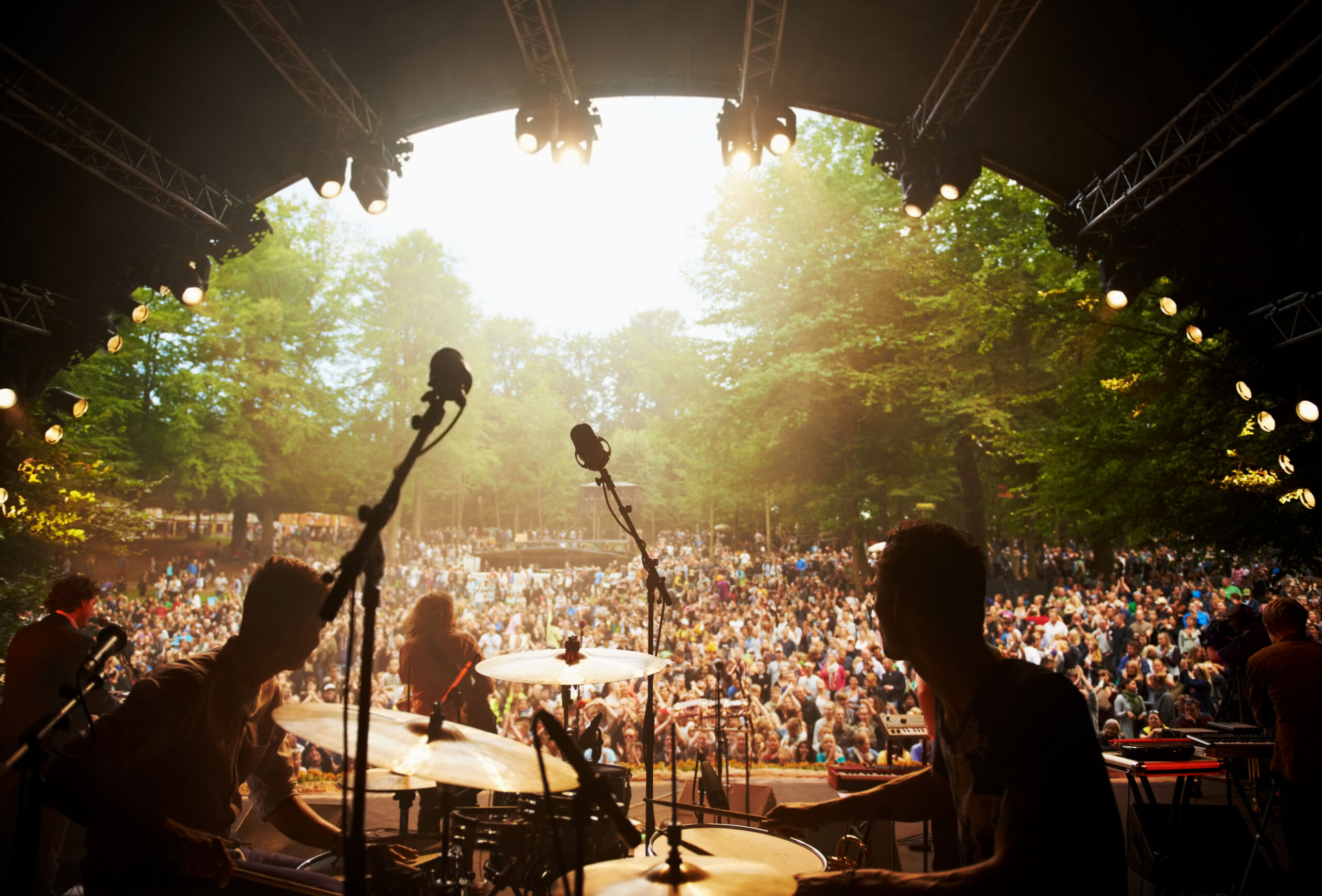 Photograph of a live outdoor concert showing band members performing on stage with musical instruments and microphones, facing a large audience seated among trees. Stage lighting and natural daylight illuminate the scene, highlighting the crowd's engagement and the event's vibrant atmosphere.
