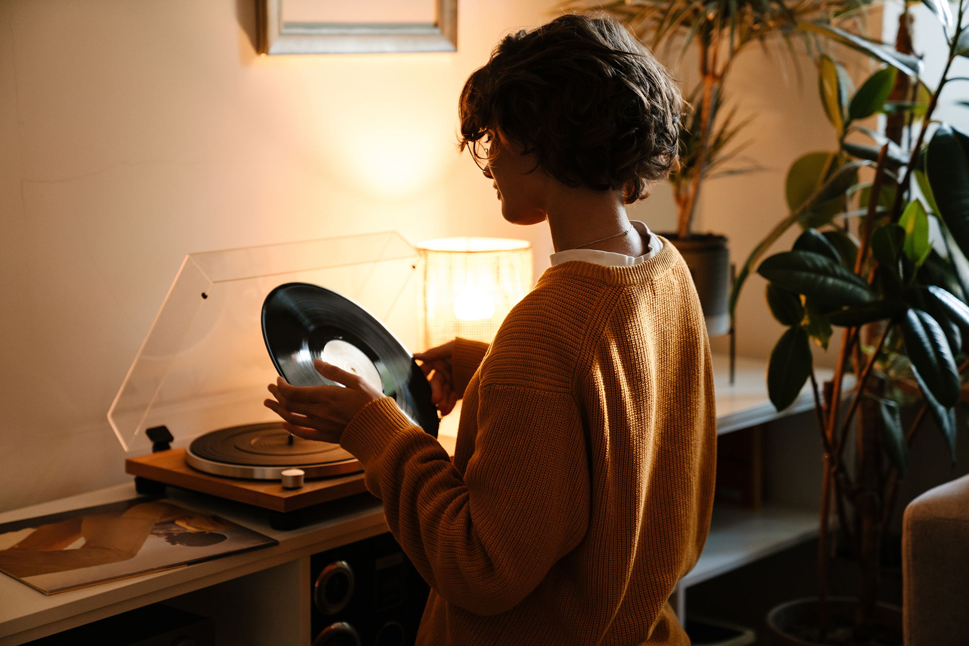 Photograph of a person placing a vinyl record onto a turntable in a warmly lit room. The scene includes a wooden shelf with audio equipment, a lamp, and several green plants, creating a cozy atmosphere.