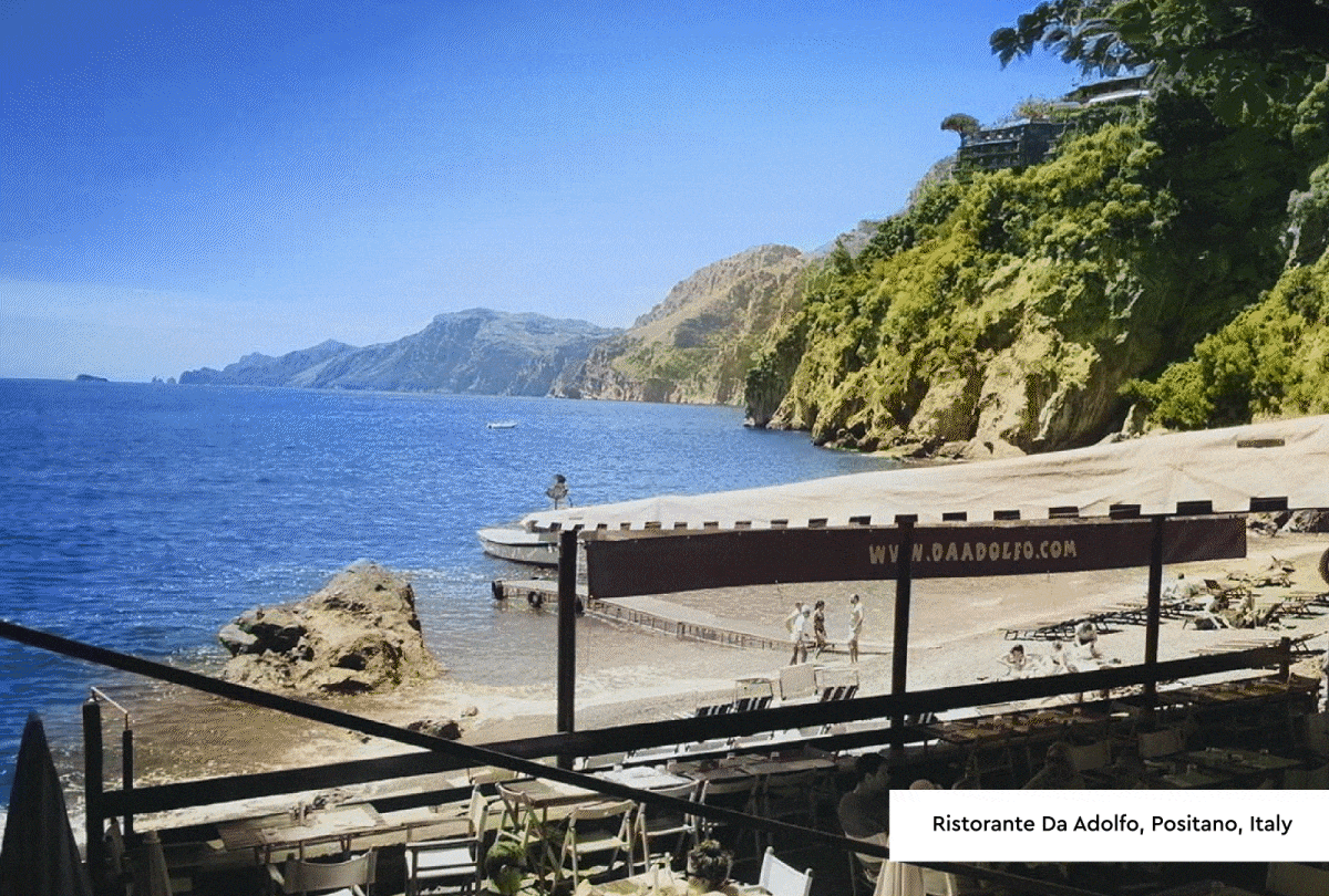 Photo 1: Cornwall -Photograph of a seaside terrace with striped awnings and lounge chairs overlooking calm blue water under a partly cloudy sky. A person is seated on a lounge chair, with a stone building and greenery visible along the waterfront, creating a peaceful coastal atmosphere. Photo 2: Positano, Italy - Photograph of a coastal scene showing a calm blue sea, rocky cliffs, and a sandy beach with a few people walking and swimming. Foreground features a shaded outdoor seating area with tables and chairs, framed by trees and a wooden structure.