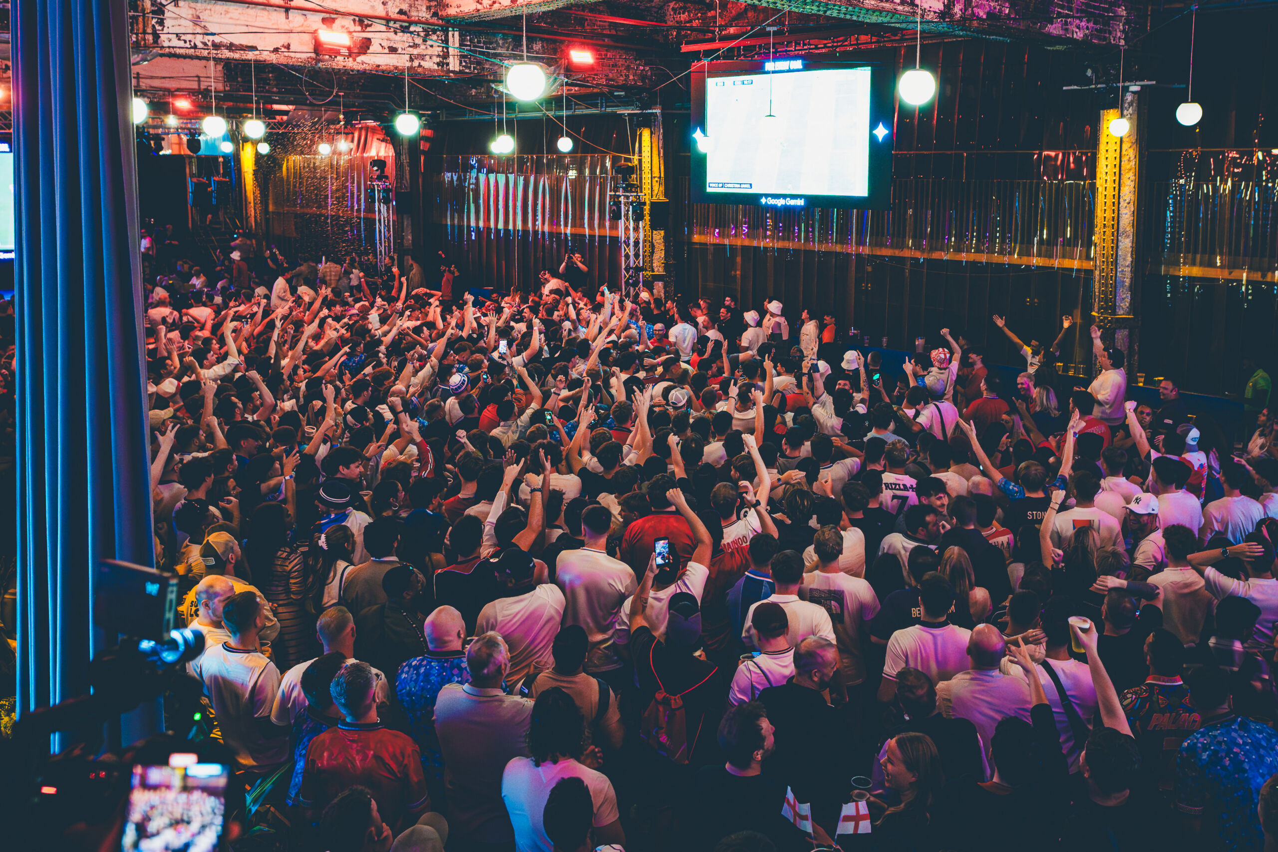 Photograph of a large crowd gathered indoors, facing a screen displaying a sports event, likely a football match. The scene features vibrant lighting with red and blue hues, and many people are raising their hands or capturing the moment with phones, indicating excitement and engagement.