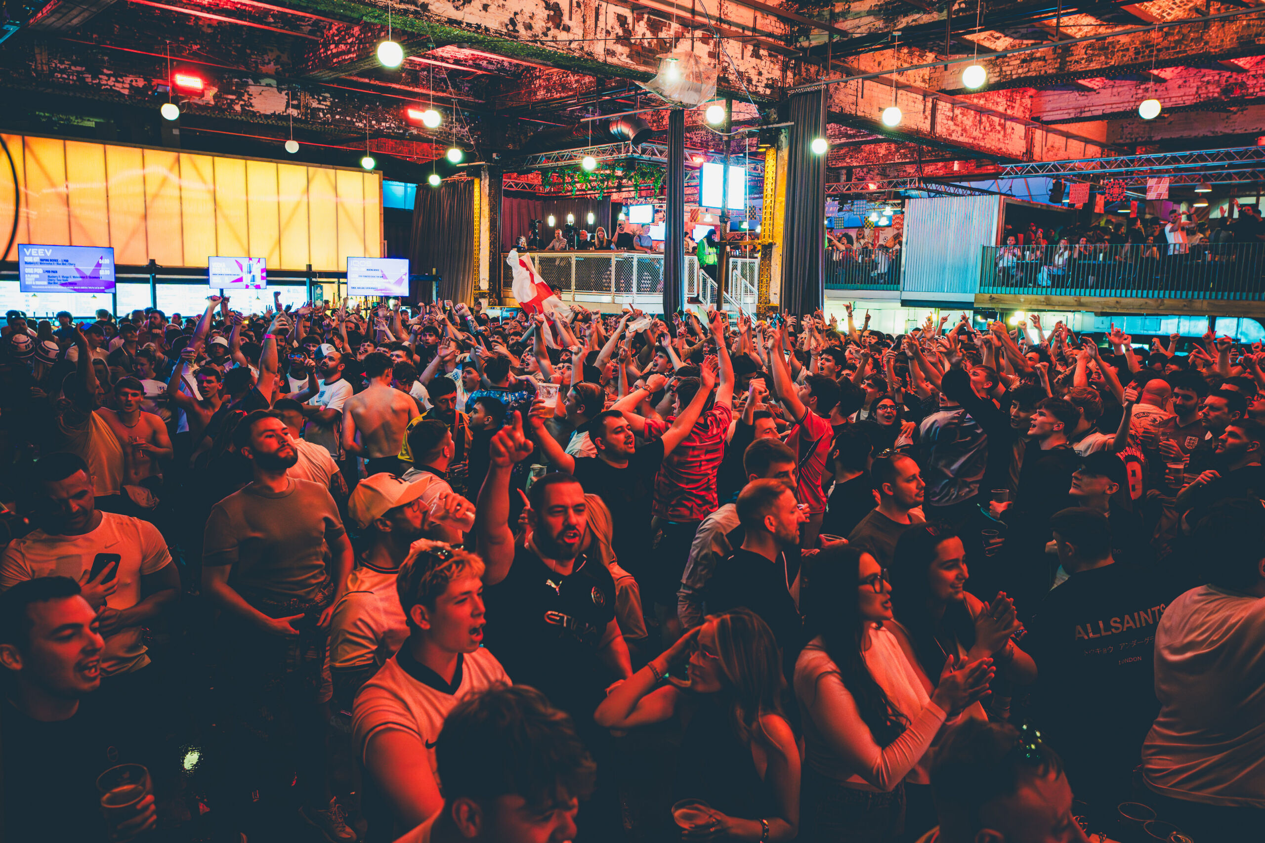 Outdoor night-time photograph of a large crowd gathered in an urban venue with string lights and a large screen displaying content. Modern buildings with illuminated windows and artistic facades form the backdrop, creating a lively atmosphere with people engaged and watching the event.
