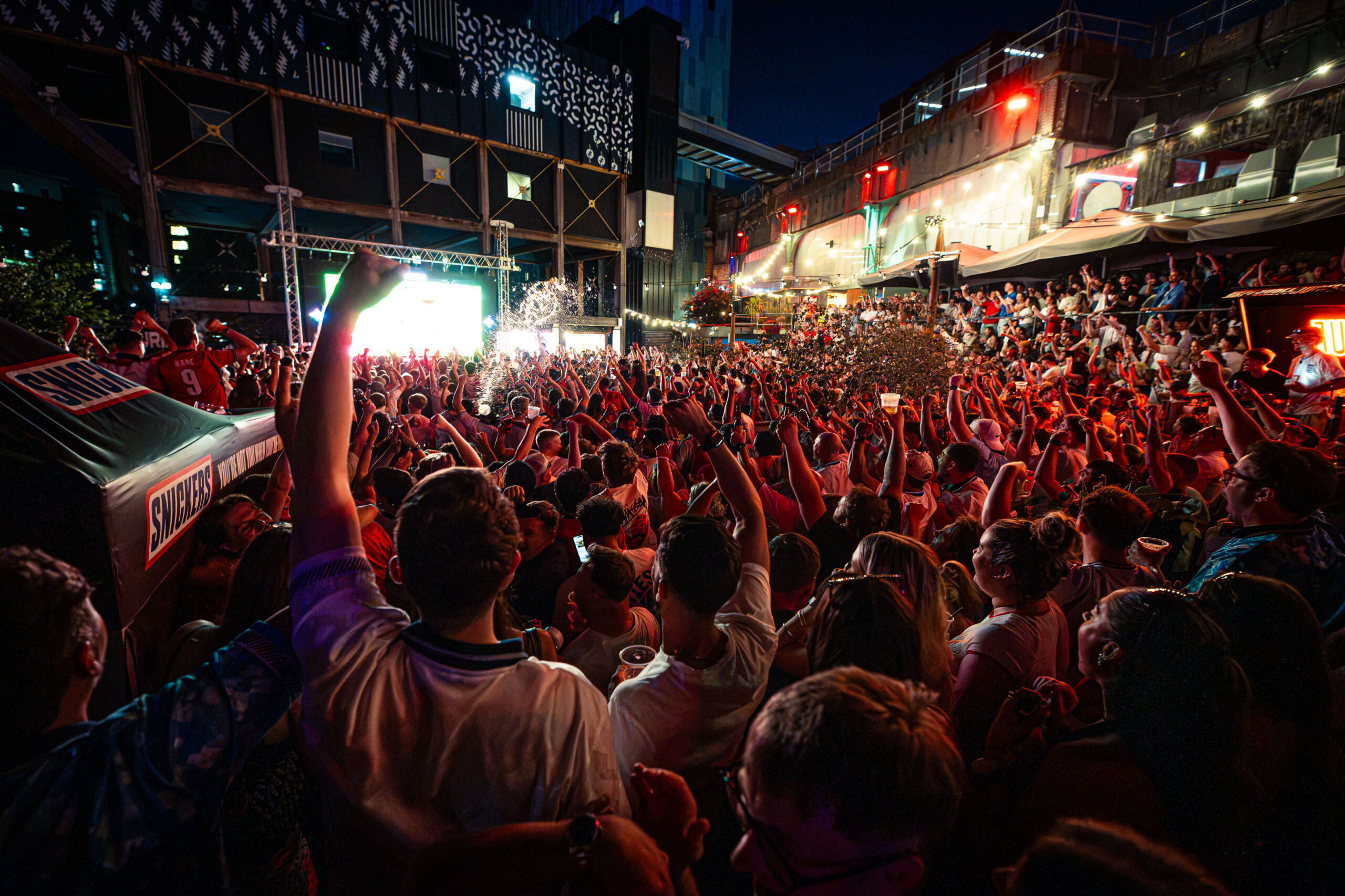 Photograph of a large outdoor crowd gathered at night, watching a brightly lit screen with many people raising their hands in excitement. The scene features vibrant lighting, confetti in the air, and a densely packed audience, suggesting a lively event or celebration.