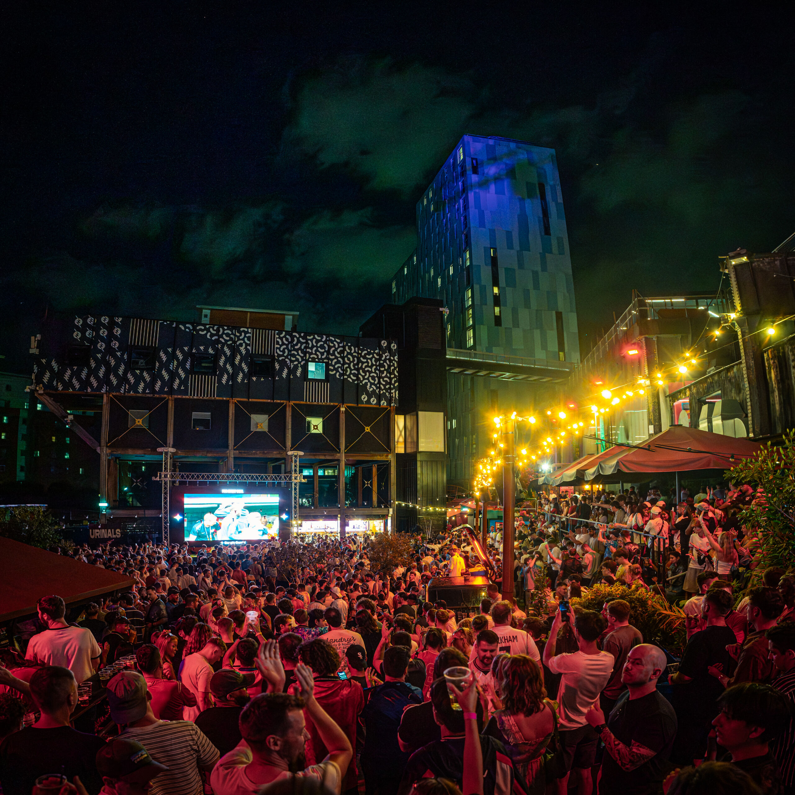 Outdoor night-time photograph of a large crowd gathered in an urban venue with string lights and a large screen displaying content. Modern buildings with illuminated windows and artistic facades form the backdrop, creating a lively atmosphere with people engaged and watching the event.