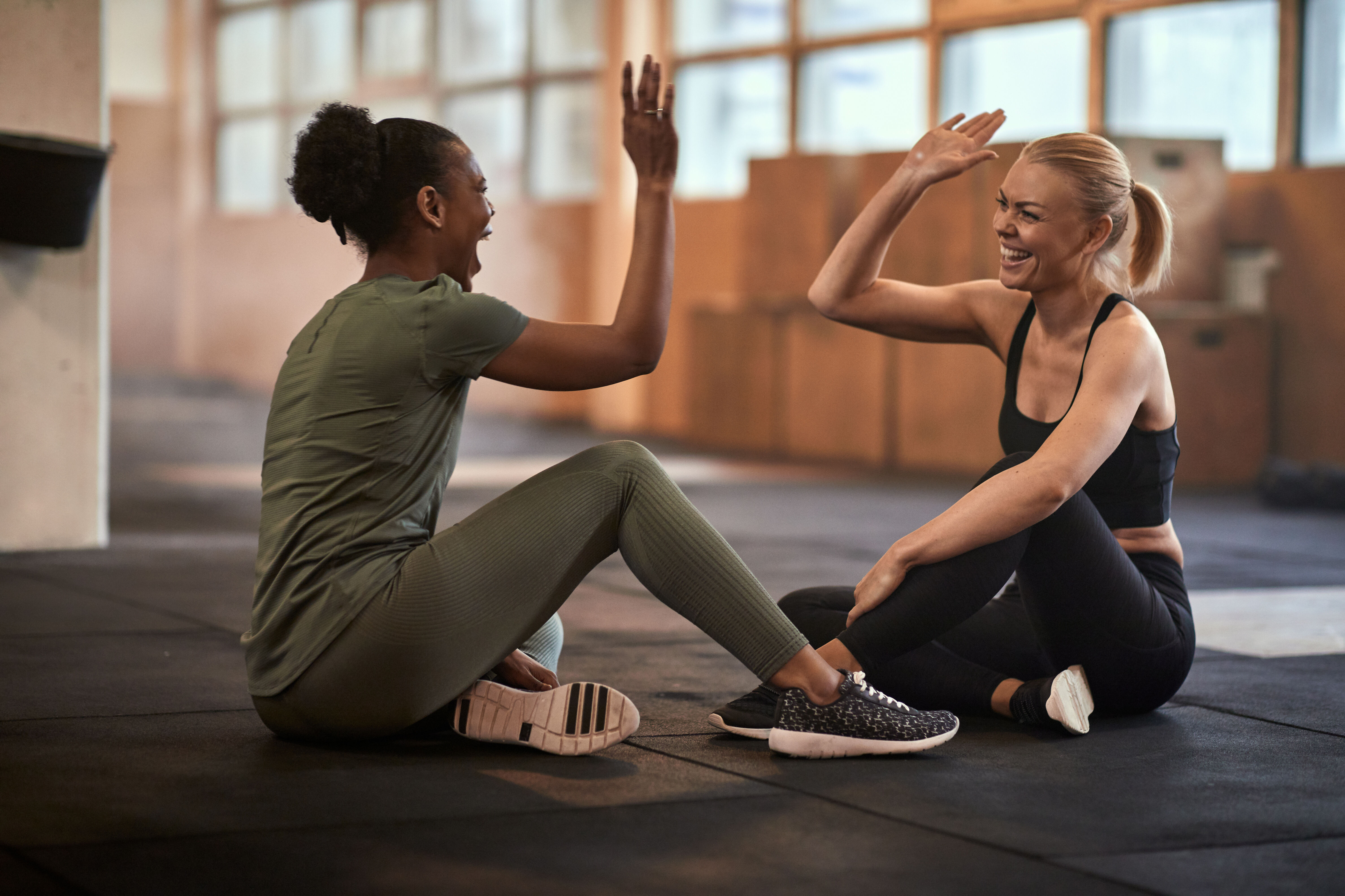 Two young women laughing and high-fiving together while sitting on the gym floor after a workout