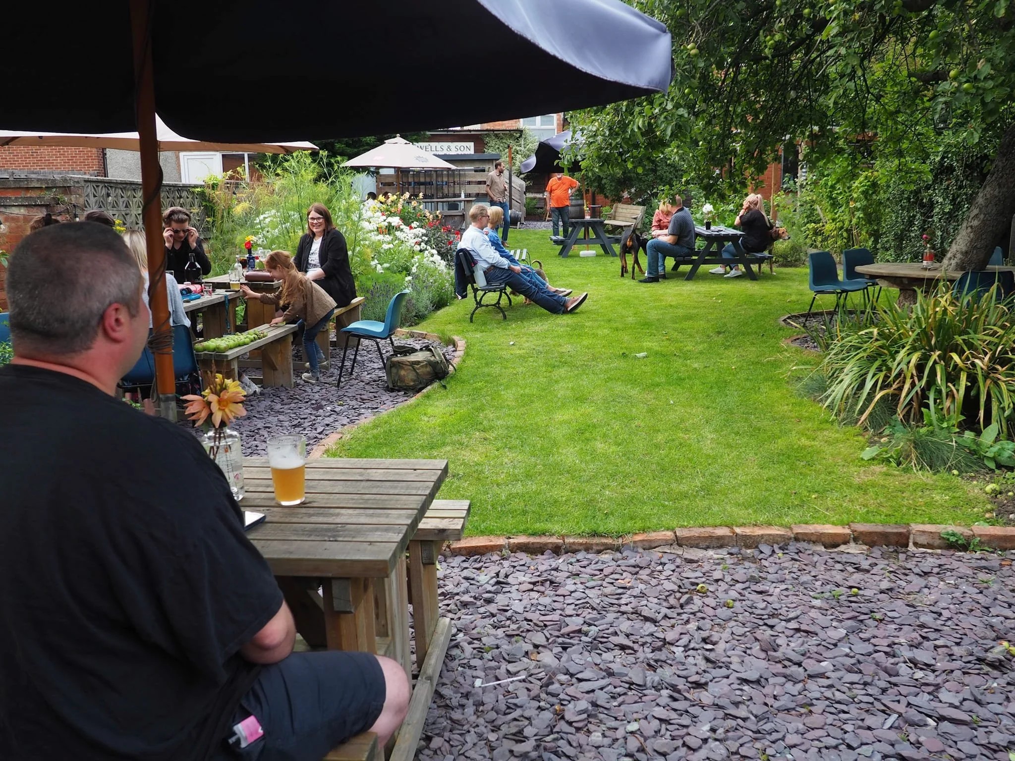 Photograph of an outdoor garden pub area with several groups of people sitting at wooden picnic tables and benches on grass and stone-paved sections. The scene includes green trees and plants, a man in the foreground with a glass of beer, and a relaxed social atmosphere on a cloudy day.