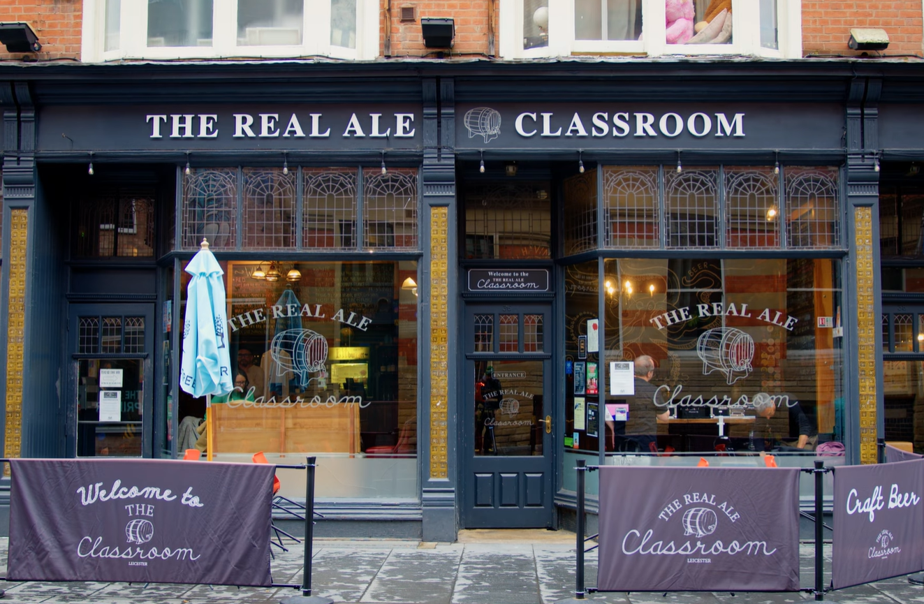 Photograph of a pub storefront named "The Real Ale Classroom" with dark blue exterior, large windows displaying the pub's logo, and outdoor seating barriers featuring welcoming text. The scene highlights a cozy, inviting atmosphere focused on craft beer education or tasting.