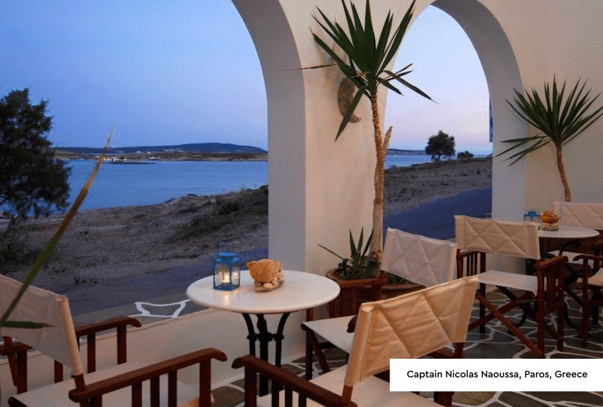 Photo 1: Whitstable - Photograph of a coastal view taken from a balcony featuring a white metal table and two chairs, with a bowl on the table. The scene includes a calm sea, a grassy promenade, a wooden utility pole, and houses along the shoreline under a clear sky at sunset. Photo 2: Paros, Greece - Photograph of an outdoor seating area at a coastal café during twilight, featuring white round tables, wooden chairs with cream cushions, and potted plants under white arches. Calm sea and distant land are visible beyond a sandy shore, creating a peaceful and scenic atmosphere.