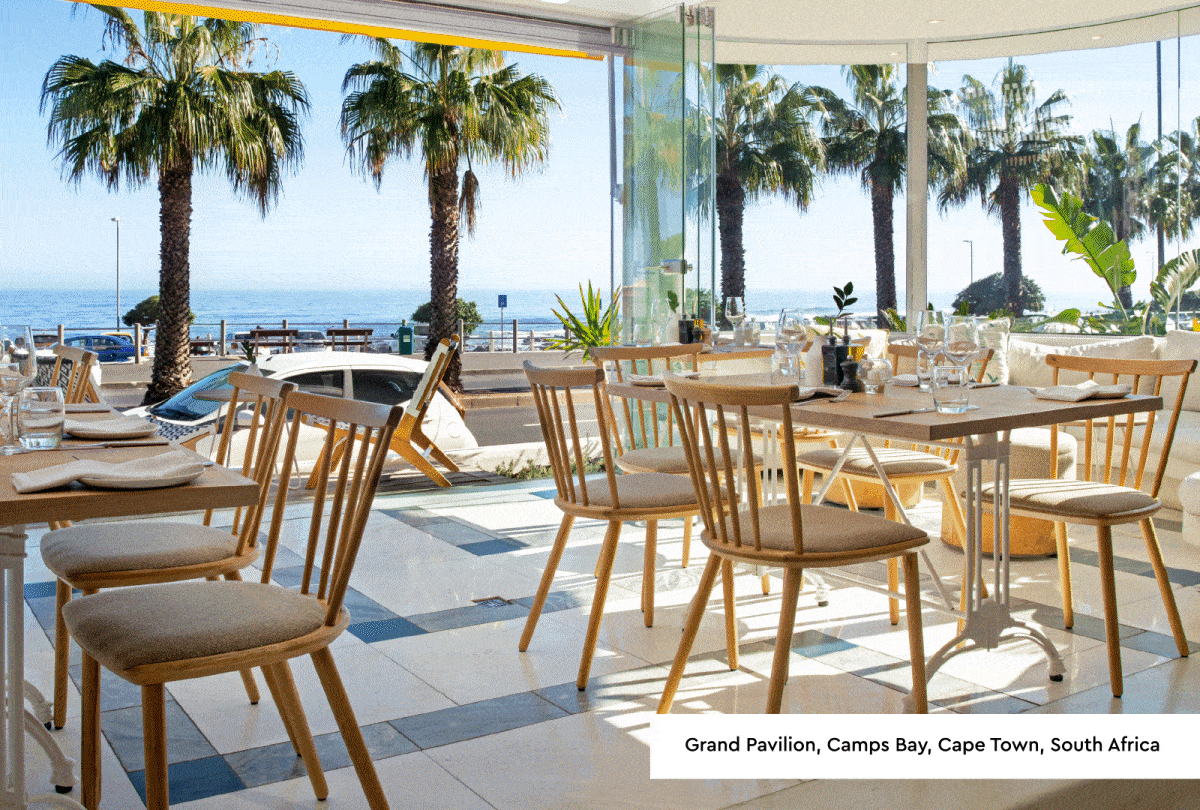 Photo 1: Worthing, Sussex: Photograph of a modern restaurant interior featuring wooden chairs, tables set with glassware, and large windows offering a sea view. Natural light highlights green plants and a clean, minimalist design, creating an inviting dining atmosphere. Photo 2: Cape Town, South Africa:Photograph of a bright, open-air seaside restaurant with wooden chairs and tables set for dining, featuring glassware and plates. Large glass windows offer views of palm trees and ocean under a clear blue sky, creating a relaxed, tropical atmosphere.