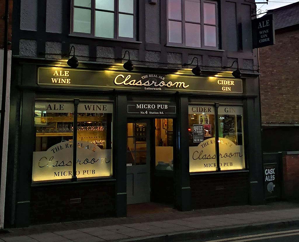 Photograph of a micro pub storefront named "Classroom" located on Station Road, featuring illuminated signage advertising ale, wine, cider, and gin. The pub has large windows with frosted glass displaying the name and type of establishment, with warm interior lighting visible through the entrance and windows.